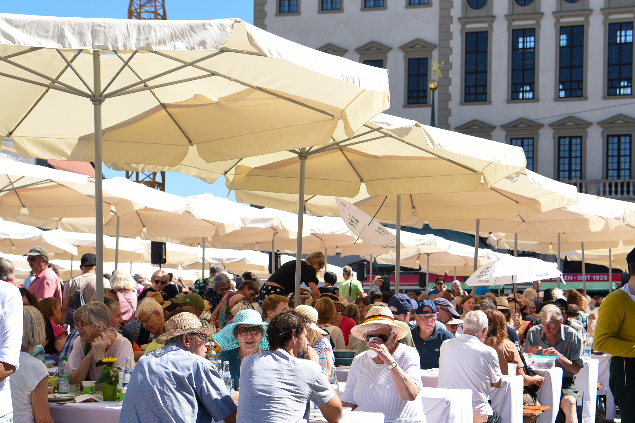 Foto der Augsburger Friedenstafel auf dem Rathausplatz. Viele Menschen sitzen an Tischen unter Sonnenschirmen beisammen.