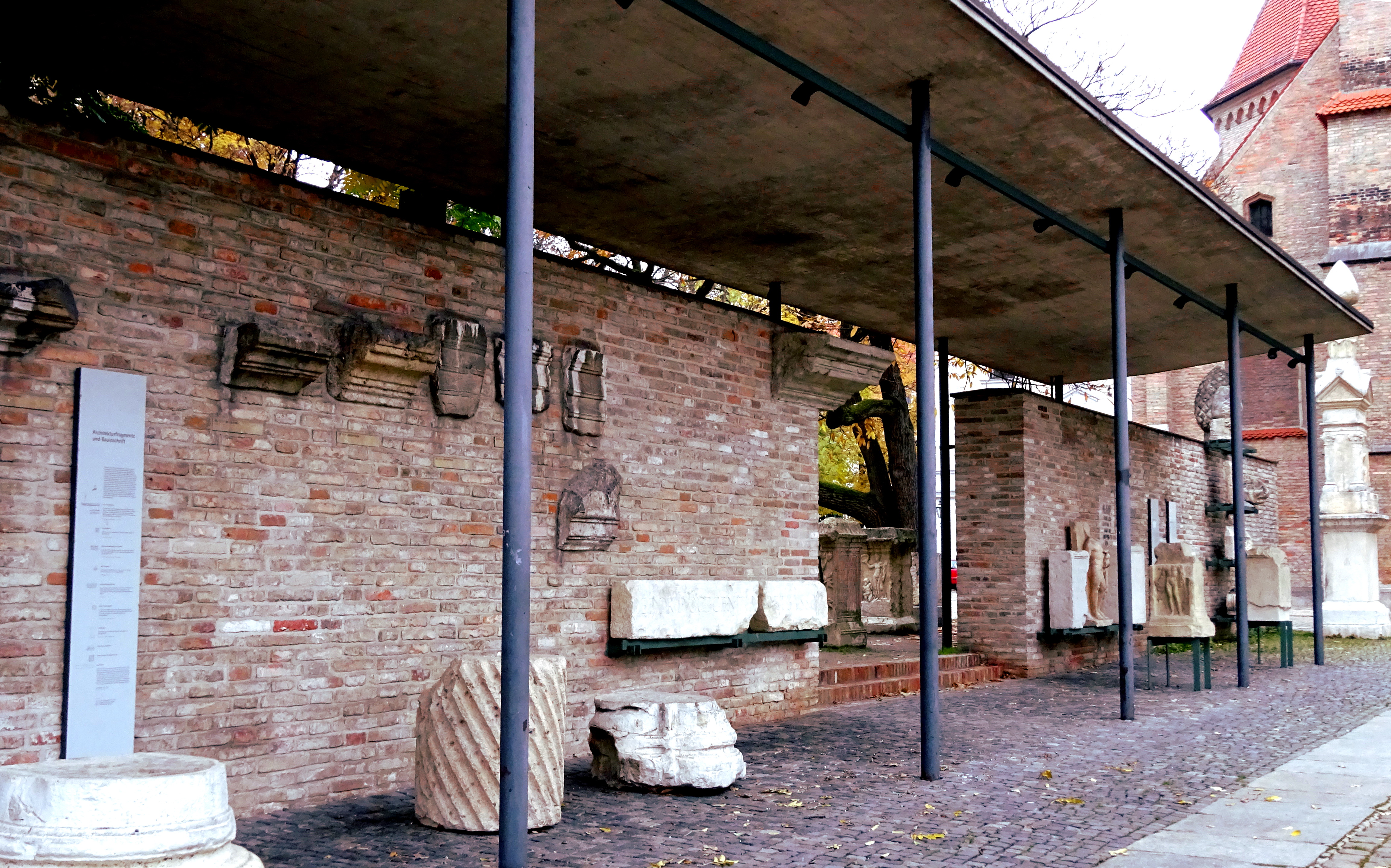 Das Bild zeigt einen überdachten teil einer Mauer, an der unterschiedliche Artefakte ausgestellt sind, die aus der Zeit der Römer in Augsburg stammen und hier ausgegraben wurden. Bild von Franz Häussler