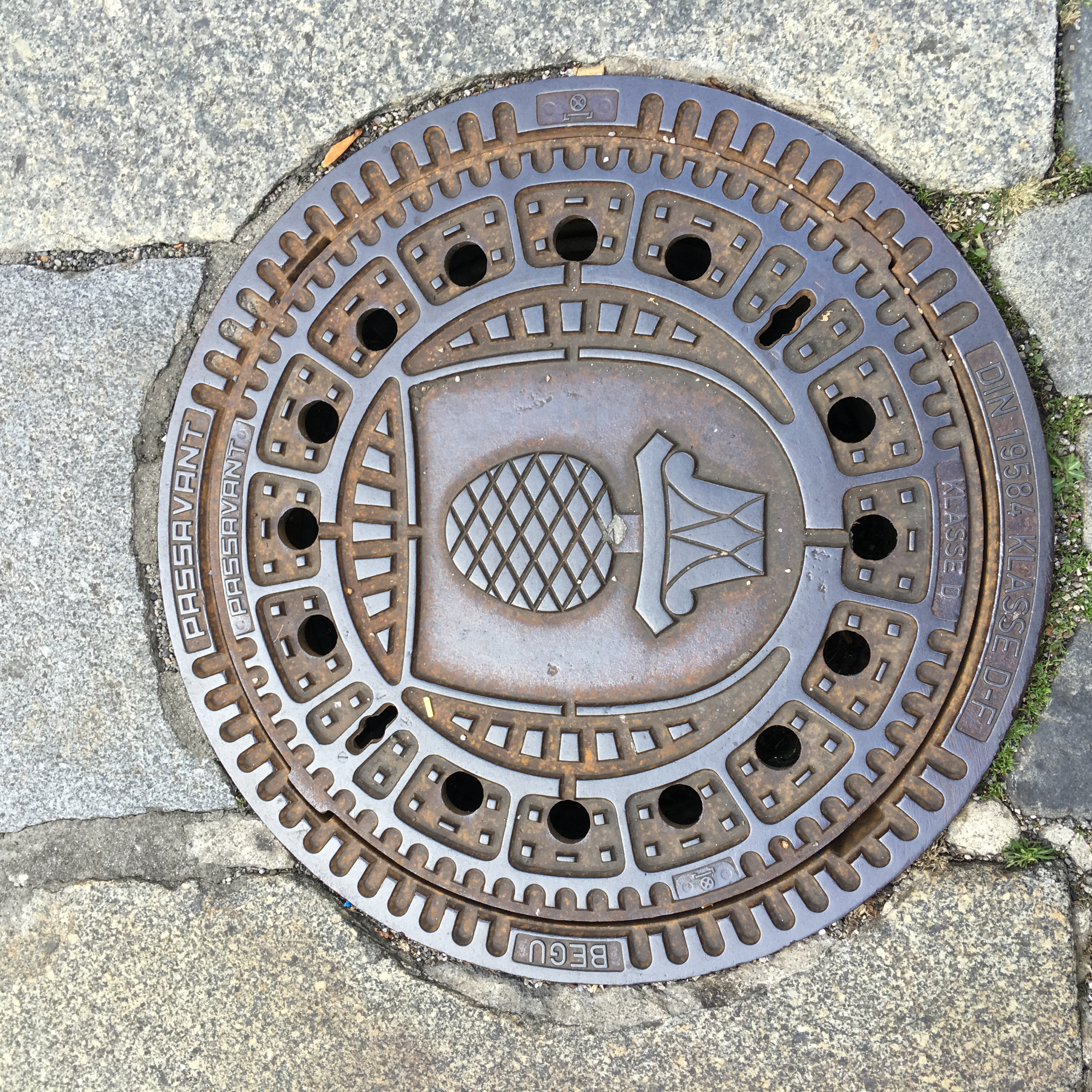 Das Bild zeigt einen Kanaldeckel der Stadt Augsburg mit der Zirbelnuss in der Mitte. Foto Katrin Jacob