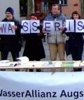 Personen der Wasserallianz Augsburg stehen mit Schildern "Wasser ist keine Ware" vor einem Tisch der Wasserallianz Augsburg