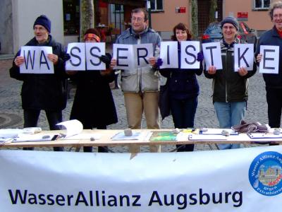 Personen der Wasserallianz Augsburg stehen mit Schildern "Wasser ist keine Ware" vor einem Tisch der Wasserallianz Augsburg