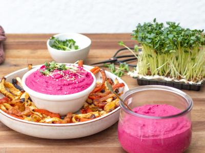 Der rote Bete Humus leuchtet pink in einer kleinen Schale die in einem Teller mit Pommes aus Gemüse thront und im Glas. Grüne Kresse bildet einen farblichen Kontrast dazu. Foto Oekokiste e.V. 