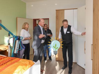 Auf dem Bild sind drei Männer, unter ihnen Landrat Martin Sailer und eine Frau bei der Besichtigung einer Musterwohnung für ein barrierearmes Zimmer zu sehen. Ein Mann öffnet eine Schranktür am rechten Bildrand und im Vordergrund steht ein Bett mit Hebevorrichtung. Das Bild stammt von Julia Pietsch