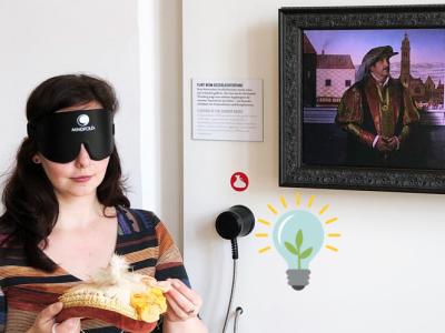 Das Bild zeigt am linken Rand eine junge Frau mit einer Dunkelbrille über den Augen, die eine gelb-orange Kappe mit Federn ertastet. Über ihr am rechten Bildrand hängt ein Gemälde, dass eine Mann und eine Frau in der Kleidung der damaligen Zeit zeigt. Bild von Regio Augsburg Tourismus GmbH_Zoltan Halmosi 