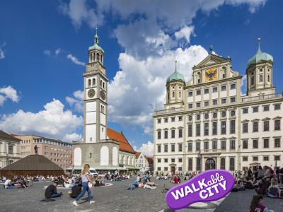 Das Bild zeigt den Rathausplatz mit Blick auf den Perlachturm und das Rathaus im Sonnenschein. Eine Frau läuft durch die Menschen, sie am Boden sitzen und die Atmosphäre genießen. Bild Norbert Liesz