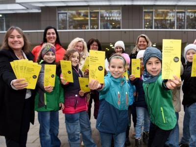 Das Foto zeigt Augsburgs zweite Bürgermeisterin Martina Wild (links) mit Kindern und Lehrkräften der Westparkschule. Sie zeigen lächelnd die gelben Flyer der Stempelkartenaktion in die Kamera. Das Foto wurde draußen vor der Schule aufgenommen.