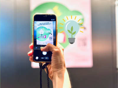 Auf dem Foto hält eine Hand ein Handy in der Hand. Auf dem Handybildschirm ist zu erkennen, dass gerade ein Foto eines "Augsburgs checkt's" Plakats gemacht wird.