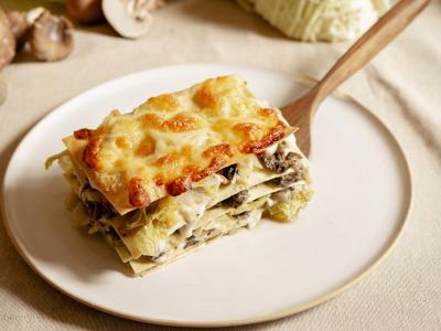 Das Foto zeigt einen hellen Teller auf dem mit einem Holzschöpfer ein Stück Wirsing-Pilz Lasagne mit Béchamelsoße serviert ist. Im Hintergrund sind Pilze und Wirsing verteilt.