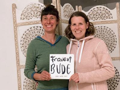 Zwei lächelnde Frauen vor einem ornamentalen Raumteiler. Die linke Frau trägt ein grün gestreiftes Shirt, die rechte einen rosa Hoodie. Sie halten gemeinsam ein Schild mit der Aufschrift "frauen BUDE – good vibes only".