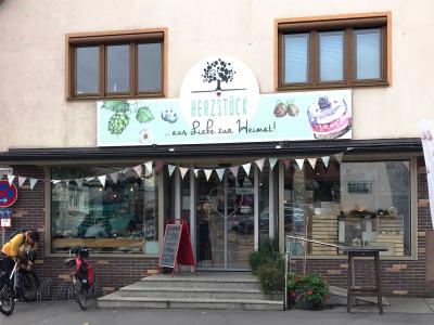Außenansicht des Herzstück Diedorf mit großer Glasfront, darüber ein Schild mit dem Text "HERZSTÜCK ... aus Liebe zur Heimat!" und dem charakteristischen Baum-Logo, vor dem Laden steht eine Person mit Lastenrad