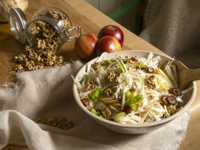 Das Bild zeigt eine Schüssel mit einem Waldorfsalat aus geraspelten Äpfeln, weissem Knollensellerie und Stücken von grünem Staudensellerie. Darauf sind halbe Walnüsse und Dattelstücke verstreut. Die Schüssel steht auf einem Holztisch auf dem außerdem ein Leinentuch, ein umgekipptes Glas Walnüsse und Äpfel zu sehen sind. Foto: Oekokiste e.V.