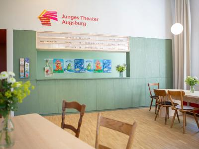Theaterfoyer mit mintgrüner Theke, Holztischen, Stühlen und Logo "Junges Theater Augsburg" an der Wand