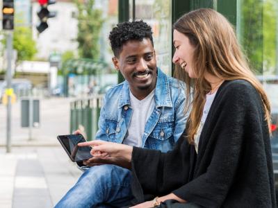 Einen junge Frau und ein junger Mann sitzen an der Haltestelle und schauen auf ein Handy. Foto Tür an Tür – Digitalfabrik gGmbH