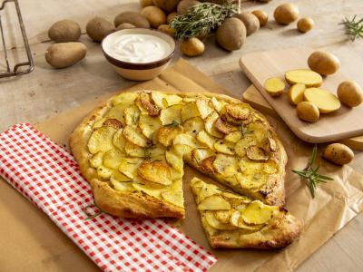 Das Bild zeigt ein eine Pizza mit knusprigen Kartoffelscheiben auf einem Teig, bestreut mit Pfeffer und Rosmarin, die auf einer Unterlage auf einem Tisch liegt.