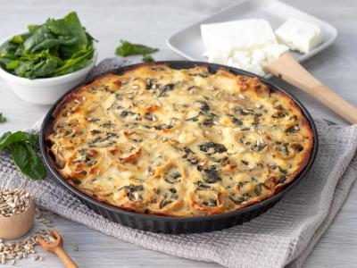 Das Bild zeigt eine flache Auflaufform mit einem goldbraun überbackenen Spinat-Pfannkuchen-Auflauf. Er steht auf einem grauen Tuch. Am linken Bildrand oben steht eine weiße kleine Schüssel, die mit Spinatblättern gefüllt ist. Am unten Rand steht eine schmale hohe Holzschüssel mit Sonnenblumenkernen darin. Am rechten oberen Rand steht ein viereckiger Teller mit einem Stück Feta darin. Bild Oekokiste e.V.