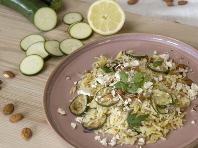 Das Bild zeigt einen Teller mit Zuchini-Zitronen-Orzo mit gerösteten Zucchinischeiben, Madelnsplittern und Flöckchen von Schafskäse bestreut. Der rosafarbene Teller steht auf einem Holzbrett auf dem links neben dem Teller Madeln, Zucchini-Scheiben und eine Zucchini sowie eine halbe Zitrone zu sehen sind. Bild Oekokiste e.V.