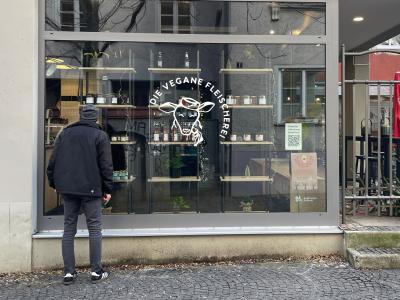 Außenansicht der Veganen Fleischerei Augsburg mit Logo im Schaufenster, Kreidetafel-Speisekarte und einem Passanten vor dem Eingang. 