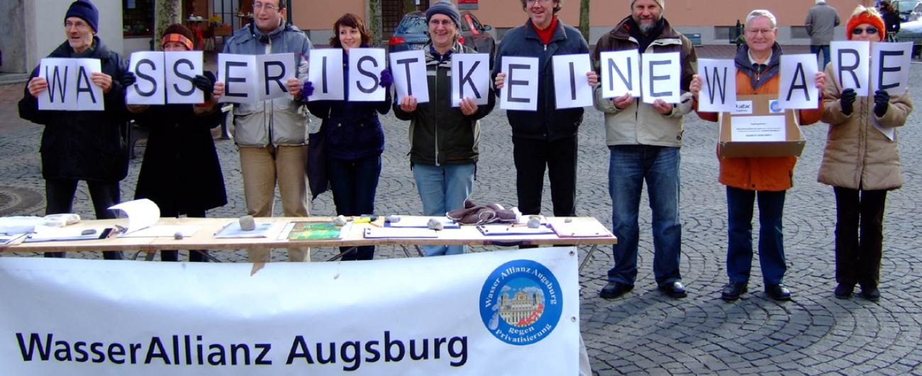 Personen der Wasserallianz Augsburg stehen mit Schildern "Wasser ist keine Ware" vor einem Tisch der Wasserallianz Augsburg