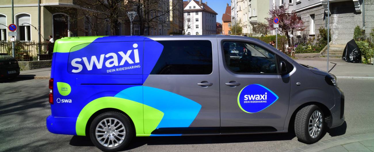 Ein silberner Van mit blau/grünen Aufklebern und dem Schriftzug swaxi steht auf einer Kreuzung im Bismarkviertel. Foto von Thomas Hosemann 