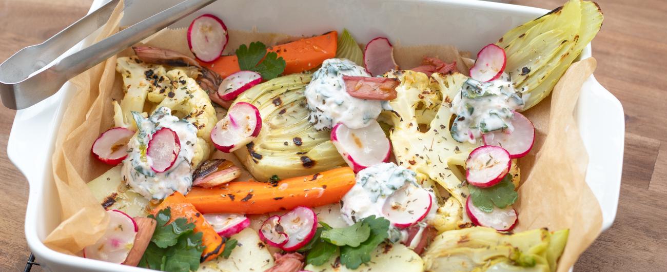 Das Bild zeigt eine bunte Mischung von gegrilltem Gemüse wie Karotten und Radieschen in einer weißen Porzellangrillschale auf einem Bett aus Backpapier. Kleine Inseln von einem Quarkdipp mit Kohlrabi- und Radieschengrün. Das Bild ist von oekokiste e.V.