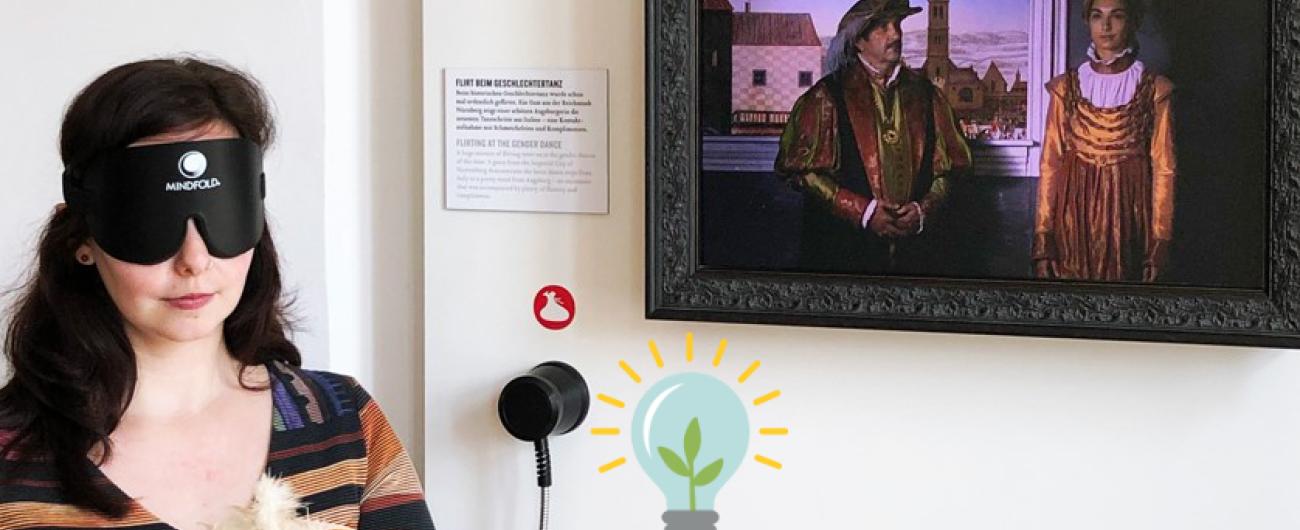 Das Bild zeigt am linken Rand eine junge Frau mit einer Dunkelbrille über den Augen, die eine gelb-orange Kappe mit Federn ertastet. Über ihr am rechten Bildrand hängt ein Gemälde, dass eine Mann und eine Frau in der Kleidung der damaligen Zeit zeigt. Bild von Regio Augsburg Tourismus GmbH_Zoltan Halmosi 