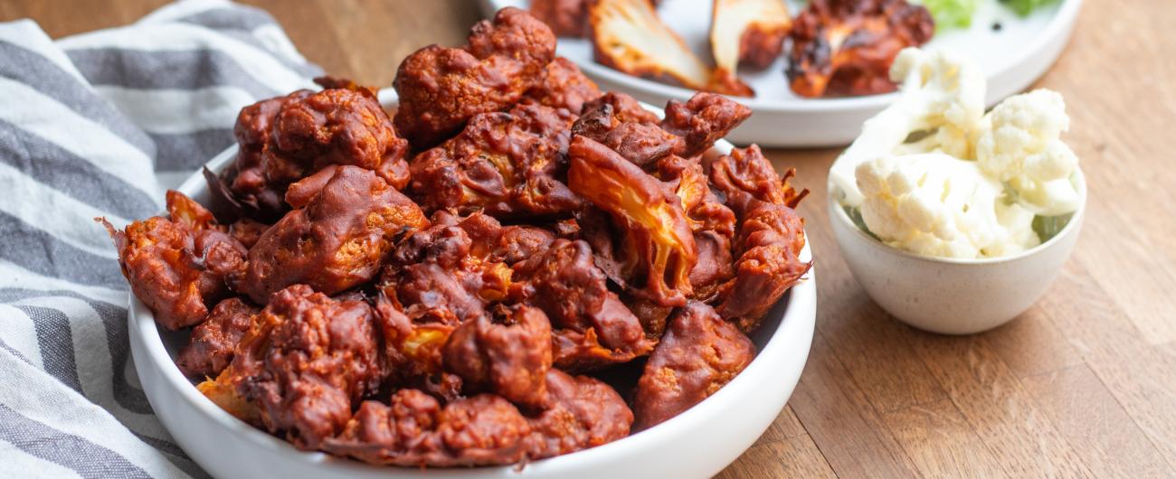 Das Bild zeigt eine kleine Schüssel mit rotbraunen Blumenkohlröschen auf einem grauweiß gestreiften Tischtuch. Daneben befindet sich eine weitere Schüssel mit Blumenkohlröschen Natur auf dem blanken Holz des Tisches. Foto Oekokiste e.V.