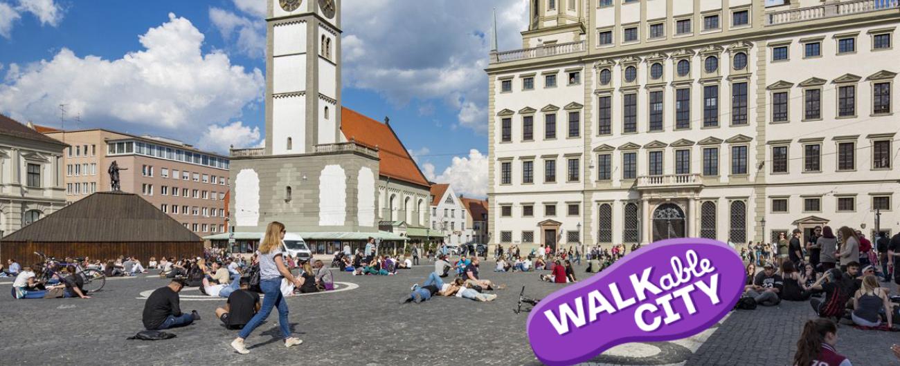 Das Bild zeigt den Rathausplatz mit Blick auf den Perlachturm und das Rathaus im Sonnenschein. Eine Frau läuft durch die Menschen, sie am Boden sitzen und die Atmosphäre genießen. Bild Norbert Liesz