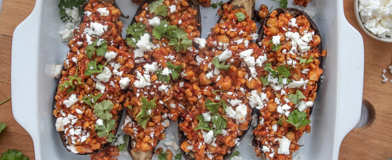 Das Bild zeigt eine weiße Auflaufform in der zwei gebackene Auberginen mit einer Füllung aus Dinkelreis zu sehen. Auf den Reis verteilt sind kleine Flocken weißen Fetas. Die Auflaufform steht auf einem Holzbrett. Bild: Oekokiste e.V.