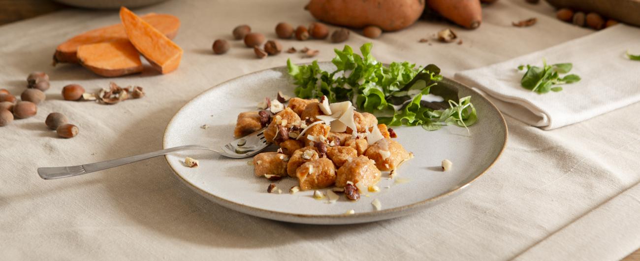 Das Bild zeigt im Vordergrund einen hellen runden Teller auf dem Süsskartoffel-Gnocchi mit grünem Salat und Parmesamstücken angerichtet sind. Am linken Rand des Tellers liegt eine Gabel. Auf dem Tisch, um den Teller, liegt eine aufgeschnittene rohe Süsskartoffel. Außerdem sind noch Haselnüsse, eine Schüssel mit Salat, eine Serviette und ein Holzteller auf dem ein Stück Parmesan liegt auf dem Tisch verteilt. Bild von oekokiste e.V.