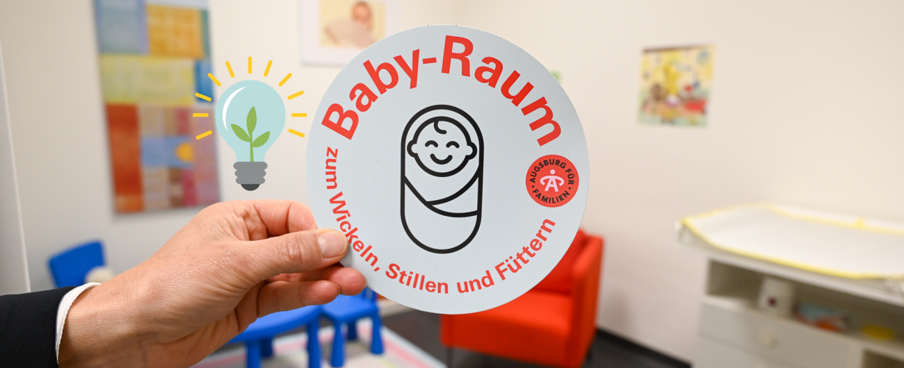 Auf dem Foto ist eine Hand zu erkennen, die einen runden Aufkleber in der Hand hält. In roten Buchstaben ist "Baby-Raum" geschrieben und passend dazu ist darunter eine schwarze Skizze eines eingewickelten lächelnden Babys gedruckt.