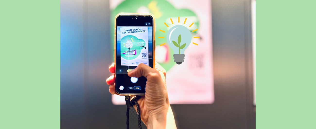 Auf dem Foto hält eine Hand ein Handy in der Hand. Auf dem Handybildschirm ist zu erkennen, dass gerade ein Foto eines "Augsburgs checkt's" Plakats gemacht wird.