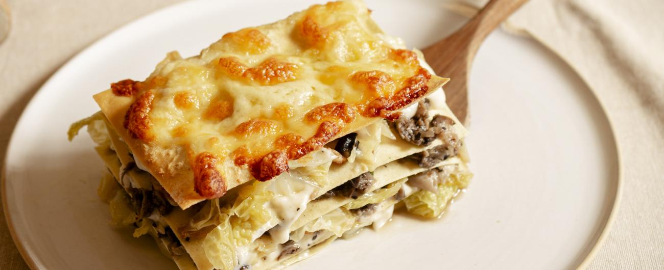 Das Foto zeigt einen hellen Teller auf dem mit einem Holzschöpfer ein Stück Wirsing-Pilz Lasagne mit Béchamelsoße serviert ist. Im Hintergrund sind Pilze und Wirsing verteilt.