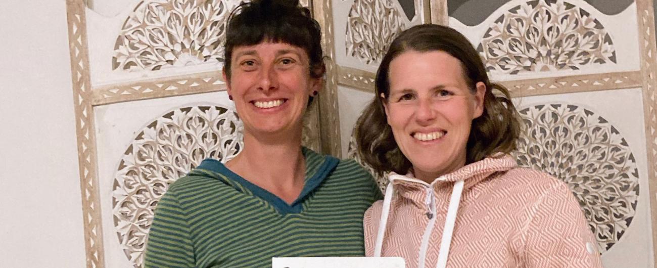 Zwei lächelnde Frauen vor einem ornamentalen Raumteiler. Die linke Frau trägt ein grün gestreiftes Shirt, die rechte einen rosa Hoodie. Sie halten gemeinsam ein Schild mit der Aufschrift "frauen BUDE – good vibes only".