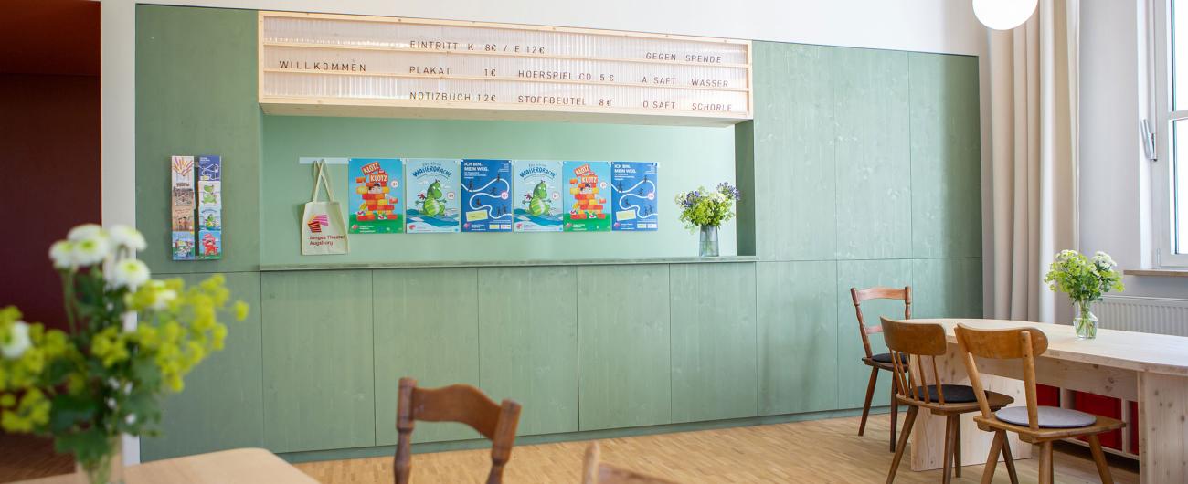Theaterfoyer mit mintgrüner Theke, Holztischen, Stühlen und Logo "Junges Theater Augsburg" an der Wand