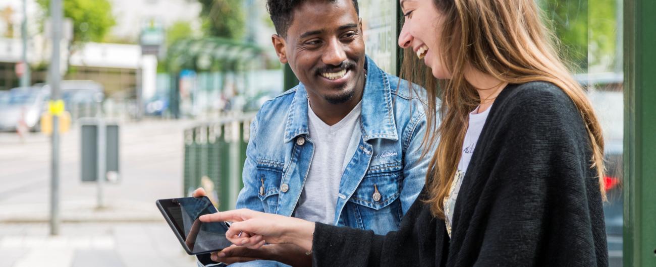 Einen junge Frau und ein junger Mann sitzen an der Haltestelle und schauen auf ein Handy. Foto Tür an Tür – Digitalfabrik gGmbH