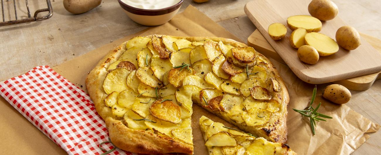 Das Bild zeigt ein eine Pizza mit knusprigen Kartoffelscheiben auf einem Teig, bestreut mit Pfeffer und Rosmarin, die auf einer Unterlage auf einem Tisch liegt.