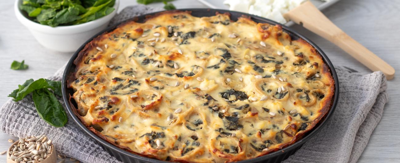 Das Bild zeigt eine flache Auflaufform mit einem goldbraun überbackenen Spinat-Pfannkuchen-Auflauf. Er steht auf einem grauen Tuch. Am linken Bildrand oben steht eine weiße kleine Schüssel, die mit Spinatblättern gefüllt ist. Am unten Rand steht eine schmale hohe Holzschüssel mit Sonnenblumenkernen darin. Am rechten oberen Rand steht ein viereckiger Teller mit einem Stück Feta darin. Bild Oekokiste e.V.