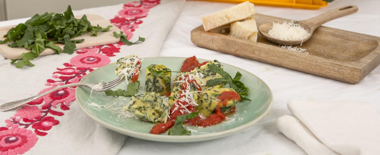 Das Bild zeigt einen Teller mit Spinat-Ricotta-Klößchen auf einem Teller mit Tomatensoße und Streifen frischen Spinats. Ein Malfatti ist auf eine Gabel gespießt. am linken Rand des mit einer rot-weißen Tischdecke gedeckten Tischs liegt ein Holzbrett mit frischen Spinat-Streifen. Am rechten Rand liegt Parmesan-Stücke in einer Holzschale. Daten auch ein Löffel mit geriebenem Parmesan. Bild Oekokiste e.V.