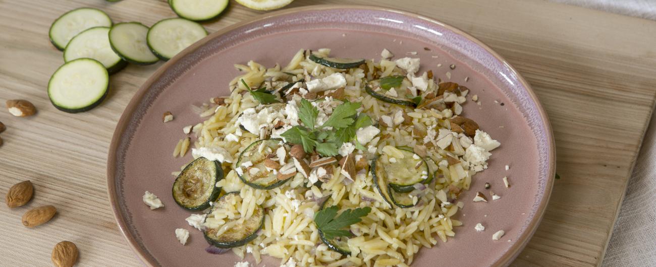 Das Bild zeigt einen Teller mit Zuchini-Zitronen-Orzo mit gerösteten Zucchinischeiben, Madelnsplittern und Flöckchen von Schafskäse bestreut. Der rosafarbene Teller steht auf einem Holzbrett auf dem links neben dem Teller Madeln, Zucchini-Scheiben und eine Zucchini sowie eine halbe Zitrone zu sehen sind. Bild Oekokiste e.V.
