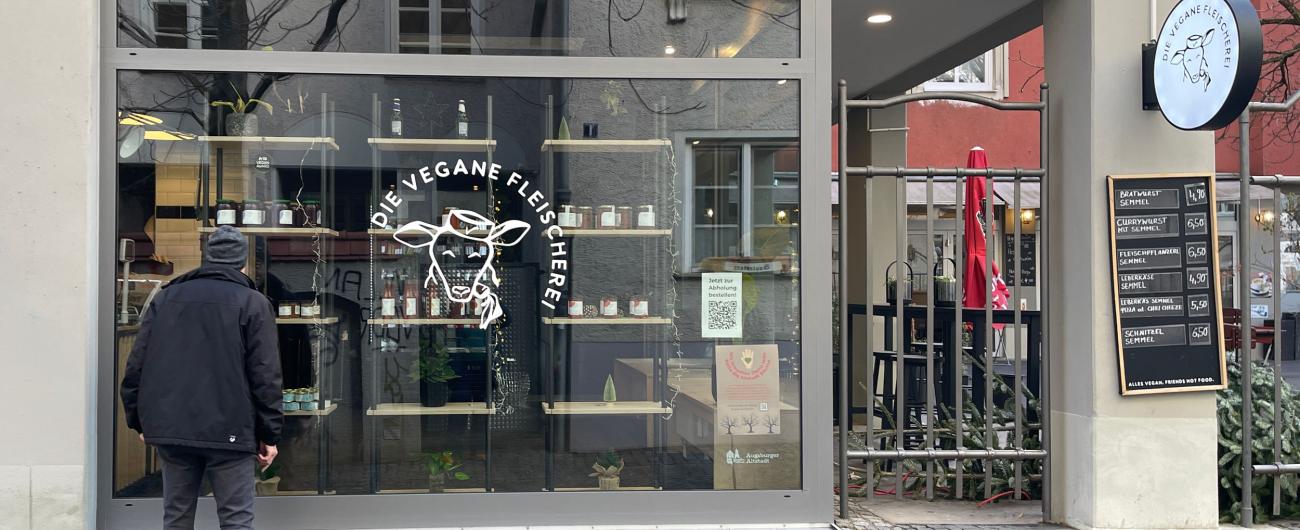 Außenansicht der Veganen Fleischerei Augsburg mit Logo im Schaufenster, Kreidetafel-Speisekarte und einem Passanten vor dem Eingang. 