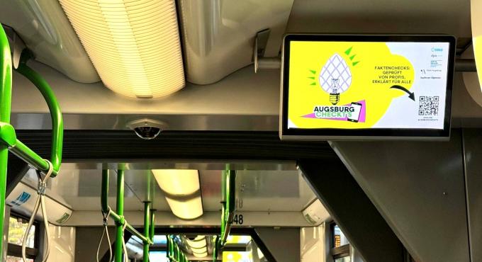 Foto wurde in einer Tram von einem Bildschirm gemacht. Auf dem Bildschirm wird gerade ein QR-Code, um Faktenchecks auf "Augsburg checkts" abzurufen, angezeigt.