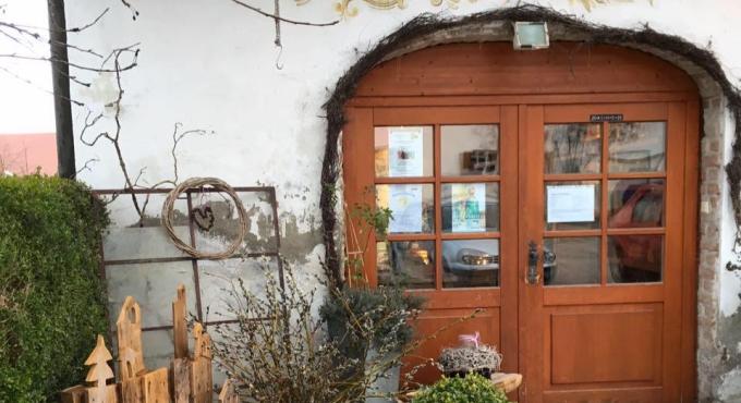 Foto zeigt den Eingang zu Höfle's Hofladen. Über der braunen Holztür mit Fenstern steht in orange-gelben Buchstaben Höfle's Hofladen. Vor dem Eingang ist ein grüner Busch zu sehen und Holzdeko.