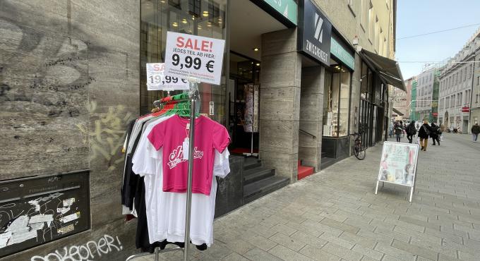 Kleiderständer mit T-Shirts vor dem Laden "Zwischenzeit" in der Augsburger Annastraße. Ein Schild zeigt "Sale! Jedes Teil ab hier: 9,99 €". Im Hintergrund die Ladenfront mit dem Schriftzug "Zwischenzeit" und der Fußgängerzone.
