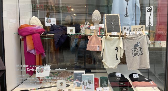 Schaufenster der Zwischenzeit in Augsburg mit bunten Schals, Babykleidung mit "Frechdachs"-Aufdruck, Strickmützen, Augsburg-Tassen und Weihnachtskarten.