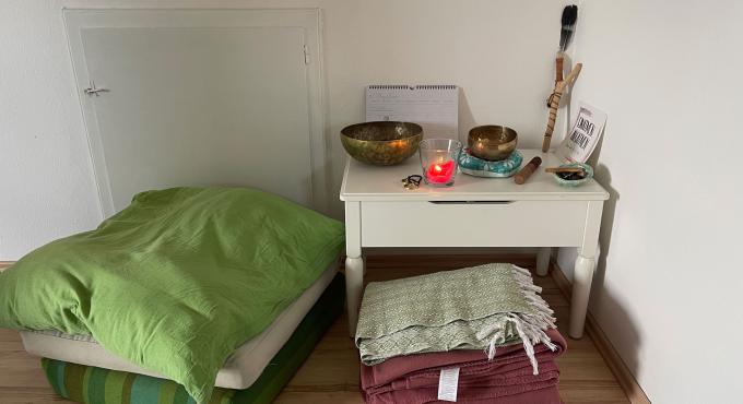 Helles Zimmer mit Einzelbett, grüner Bettwäsche und weißem Nachttisch. Darauf Klangschalen, Kerze, Räucherstäbchen und spirituelle Dekoration. Ruhiger, minimalistischer Rückzugsort.