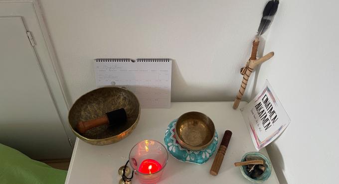 Weißer Nachttisch mit zwei Klangschalen, Klöppel, brennender Kerze, Feder, Kalender und Buch "Frauen heilen". Spirituelle Atmosphäre mit Fokus auf Achtsamkeit und Selbstfürsorge.