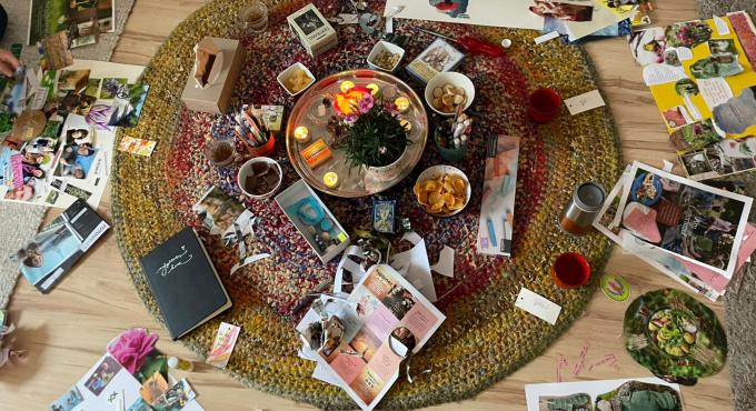 lick von oben auf einen runden, bunten Teppich, auf dem Fotos, Magazine, ein schwarzes Notizbuch, Kerzen, Snacks und Getränke verteilt liegen. Die Szene zeigt ein kreatives Treffen in der Frauenbude in Hochzoll mit gemütlicher Atmosphäre.