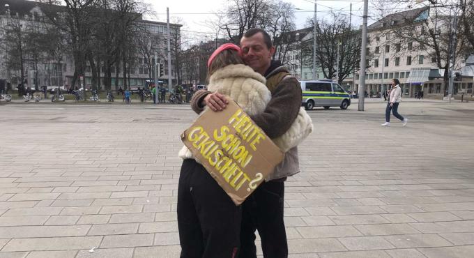 Umarmung zweier Menschen am Königsplatz in Augsburg mit einem Schild "Heute schon gekuschelt".