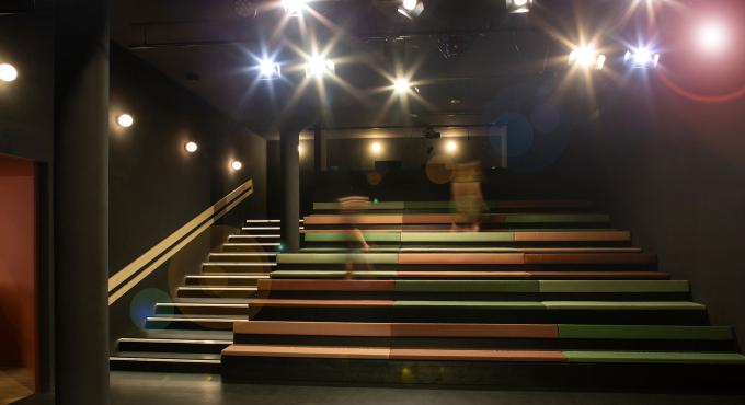 Theaterzuschauerraum mit gestaffelten Sitzbänken in Grün-, Orange- und Brauntönen, beleuchteten Stufen und Deckenscheinwerfern