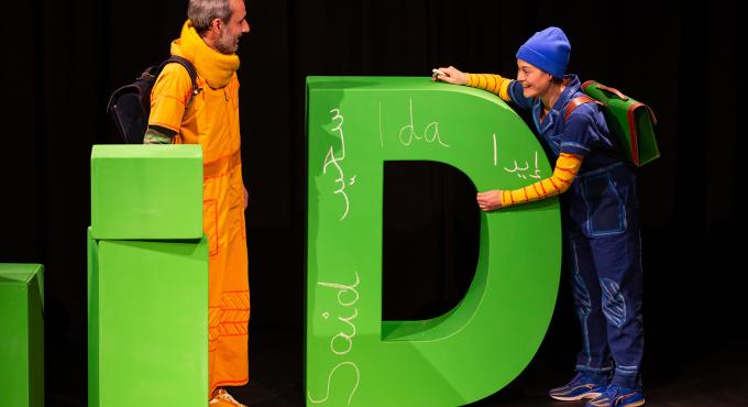 Zwei Theaterschauspieler in bunten Kostümen stehen neben großem grünem Buchstaben D auf Bühne mit mehrsprachigen Beschriftungen